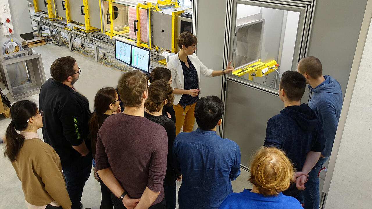 Gruppe an Personen neben Sichtfenster einer Pr&uuml;fstandskammer mit erkl&auml;nder Person vor der Gruppe